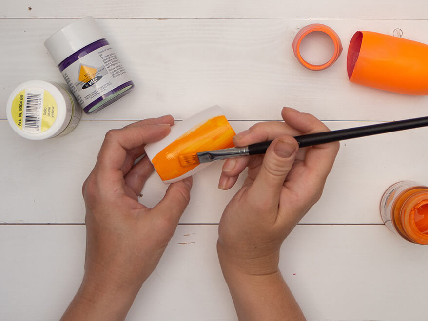 Jemand malt das Ober- und das Unterteil einer auseinandergeschnittenen, weißen Plastikflasche mit einem Pinsel mit orangefarbener Acrylfarbe von Baier und Schneider an. Der Untergrund ist hell im Hintergrund stehen weitere Farben und Flaschenteile. Foto: Johanna Rundel