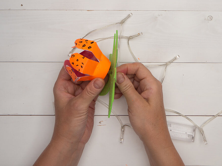 Jemand steckt Blätter aus grünem Moosgummi von Knorr Prandell, sowie eine, aus einer kleinen Plastikflasche upcycelte Blume auf ein Birnchen einer Lichterkette. Der Untergrund ist hell. Foto: Johanna Rundel