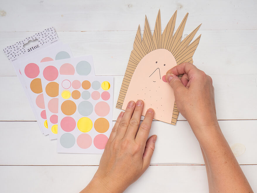 Jemand klebt zwei rosa Klebepunkte von Artoz als Bäckchen auf eine, zu einem Igel umgestalteten, braunen Papiertüte. Der Untergrund ist weiß. Foto: Johanna Rundel 
