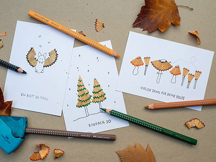 Auf einem Kraftpapier liegen drei selbstgebastelte Postkarten. Auf ihnen sind verschiedene Motive zu sehen, die aus den Abrieben vom Spitzen entstanden sind. Eine Fledermaus, zwei Tannen unter Sternen und eine Schar Waldpilze. Drumherum sind Herbstblätter, Spitzer-Reste und ein Spitzer von Faber-Castell, in dem ein Stift steckt, verteilt. Foto: Johanna Rundel