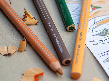 Auf einem Kraftpapier liegen viele verschiedenfarbige Buntstifte der Grip-Serie von Faber-Castell um eine geöffnete Dreifachspitzdose im Kreis. Einige Spitzreste sind dazwischen verteilt. Foto: Johanna Rundel