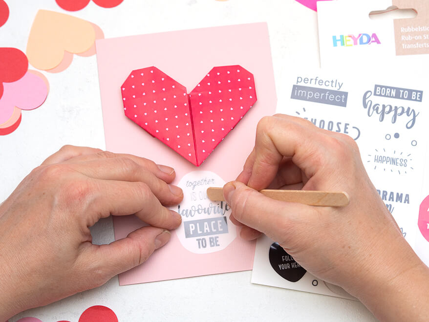 Auf einem hellen Untergrund liegt eine Valentinstagskarte, auf die eine rotes Origami-Herz aus Papier geklebt wurde. Jemand trägt darunter mit einem Holzspatel einen "Rubbelsticker" von Heyda auf. Der Schriftzug lautet: "together is our favourite place to be". Foto: Johanna Rundel