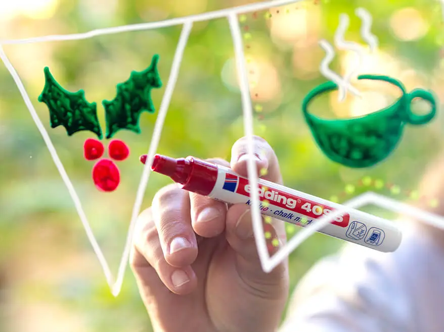 Jemand malt mit einem roten Kreidemarker von edding eine weihnachtliche Wimpelkette auf eine Fensterscheibe. Foto: Johanna Rundel