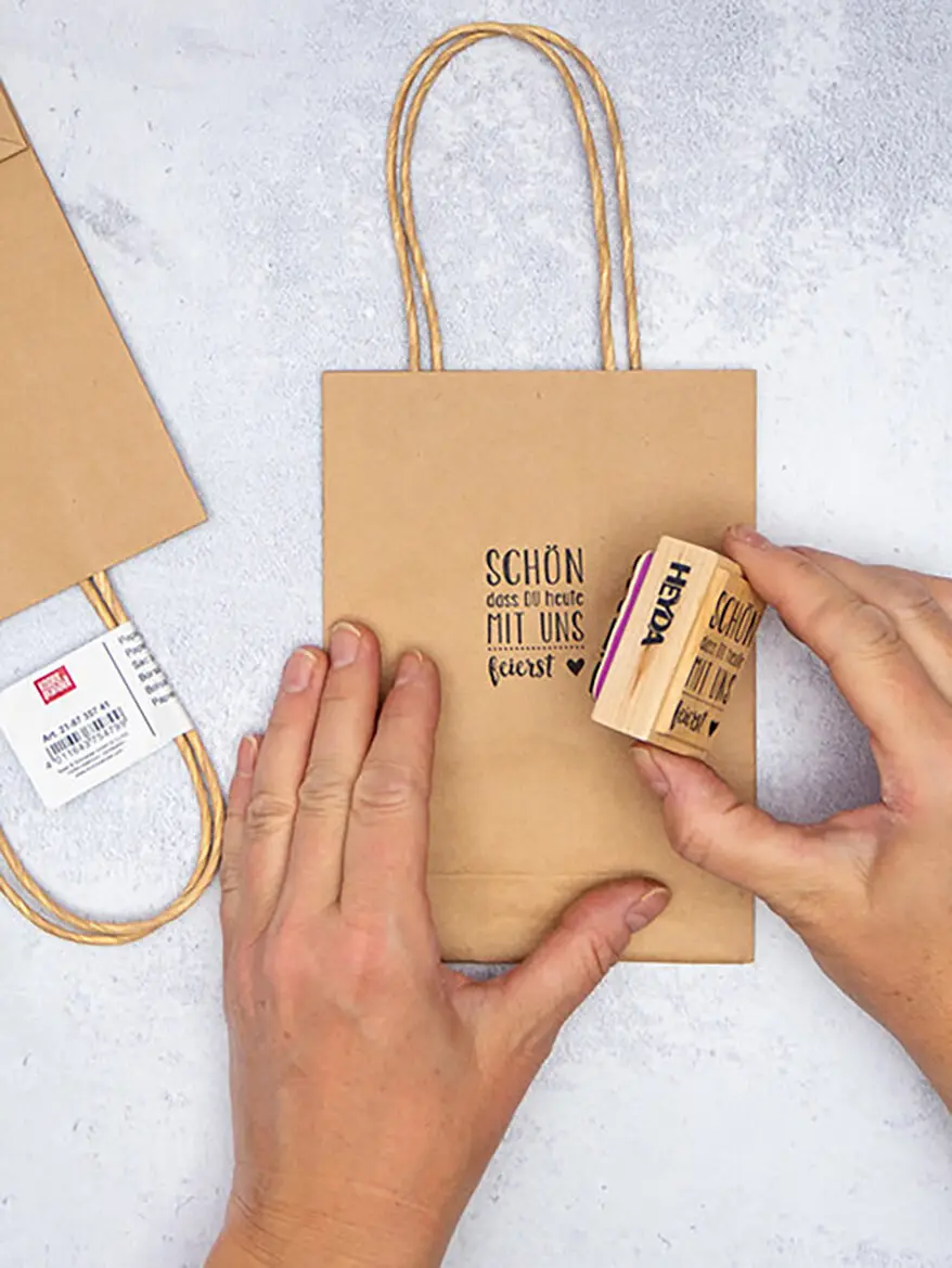 Jemand stempelt „Schön, dass du heute mit uns feierst“ mit einem Stempel von HEYDA auf eine braune Papiertüte von KNORR prandell. Foto: Johanna Rundel