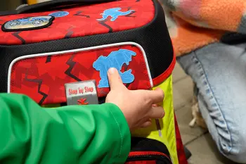 Ein Kind befestigt einen Reflektorsticker in Drachenform an einem roten Schulranzen von Step by Step.
