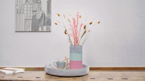 Bunte DIY-Betonvase mit Blumen steht in einer Betonschale auf einem Tisch. 