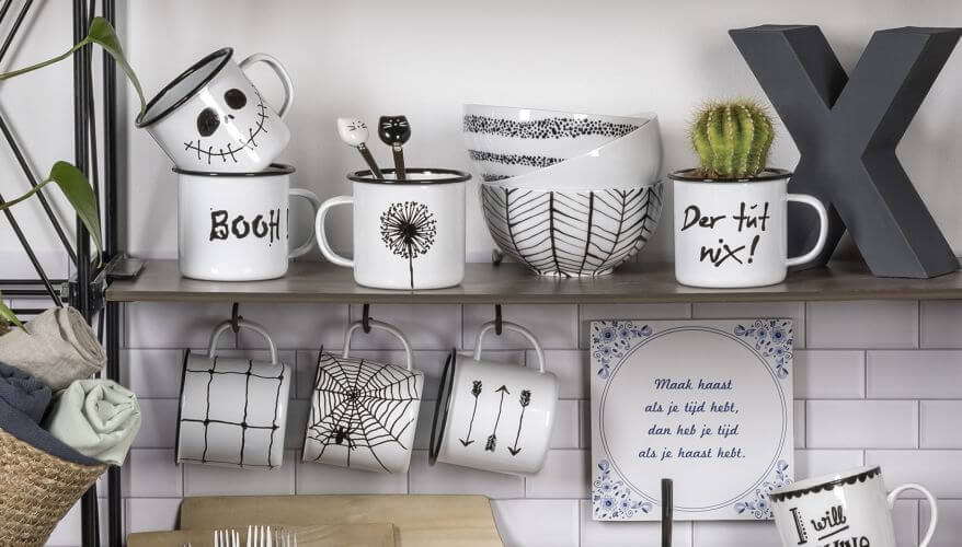 Weiße Emailletassen stehen in auf einem Küchenrgal und hängen an Haken darunter. Sie wurden mit dem Porcelain Pen von Knorr Prandell selbst bemalt und beschriftet, es sind verschiedene Motive zu sehen: Zum Beispiel Halloweenmotive, eine Pusteblume und grafisch verzierte Müslischalen.
