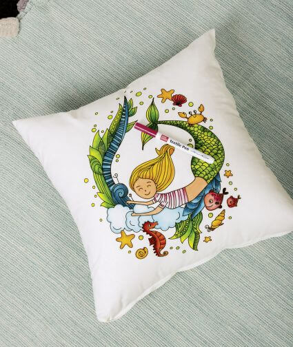 Man blickt von oben auf ein selbstbemaltes Kissen, auf dem eine Meerjungfrau und weitere maritime Motive (z.B. Fisch, Seepferdchen, Seesterne) in kindlichem Stil zu sehen sind. Das Kissen wurde mit Stoffmalstiften der Marke Knorr Prandell gestaltet.