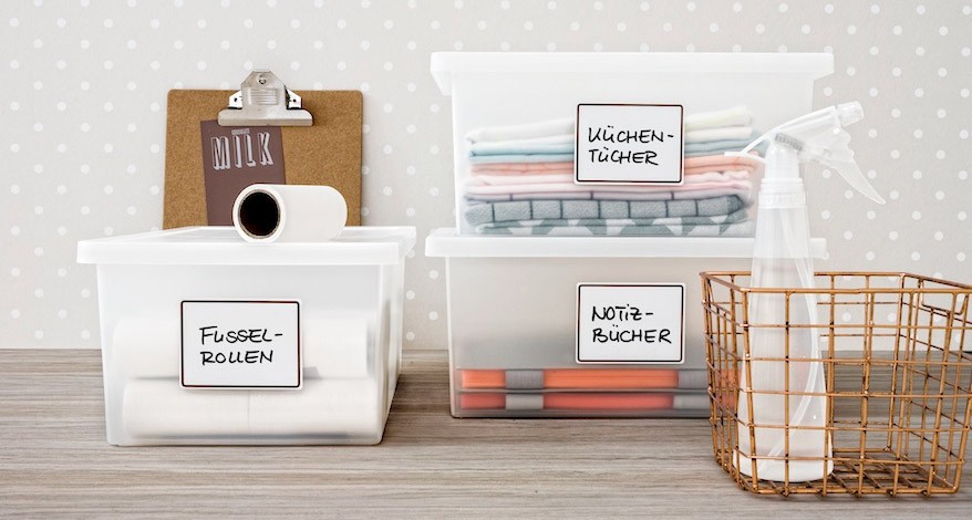 Auf einem Holztisch stehen diverse Ordnungsboxen. Diese sind mit Etiketten der Marke Avery Zweckform beklebt und beschriftet.