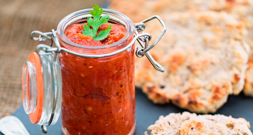Bild eines geöffneten Einmachglases, welches mit rotem Chutney gefüllt ist. Im Hintergrund ist geröstetes Fladenbrot zu sehen.