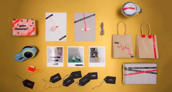 Auf einem gelben Untergrund liegen verschiedene Fotos, Geschenkanhänger, Geschenktüten und Geschenke, die mit Prägebändern von Dymo beschriftet und gestaltet wurden. Es sind außerdem zwei Prägegeräte von Dymo zu sehen.
