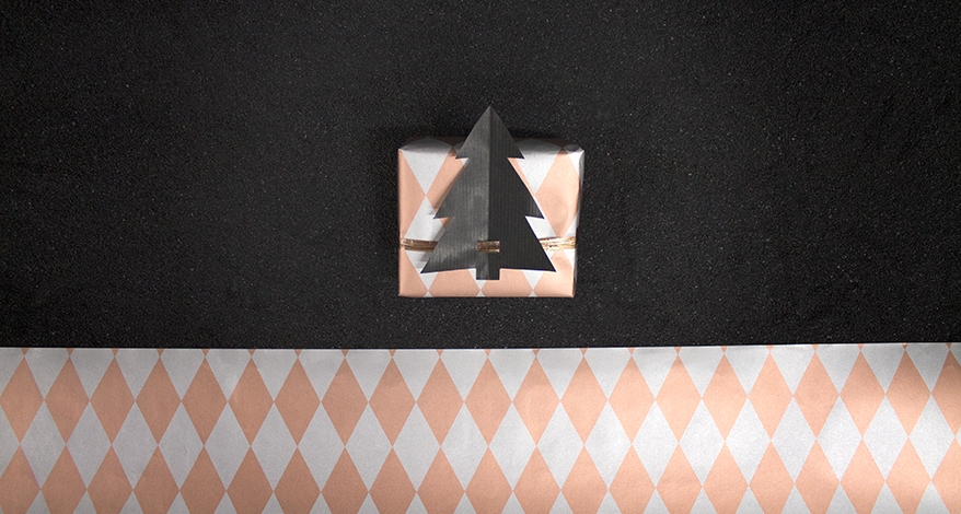 Ein quadratisches Geschenk ist in goldenes karriertes Papier eingepackt. Es ist mit einem schwarzen, aus Papier geschnittenem Tannenbaum verziert. Daneben liegt das Papier als Muster.