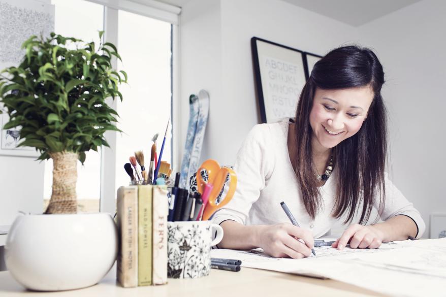 Johanna Basford sitzt an ihrem Schreibtisch und schreibt etwas auf. Neben ihr steht eine Tasse mit Stiften, Pinseln