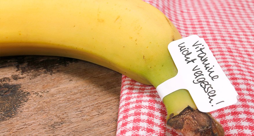 Eine Banane liegt halb auf einem Holztisch und halb auf einem rot-weiß karierten Tischtuch. An der Banane klebt ein Etikett mit der Aufschrift "Vitamine nicht vergessen!". 