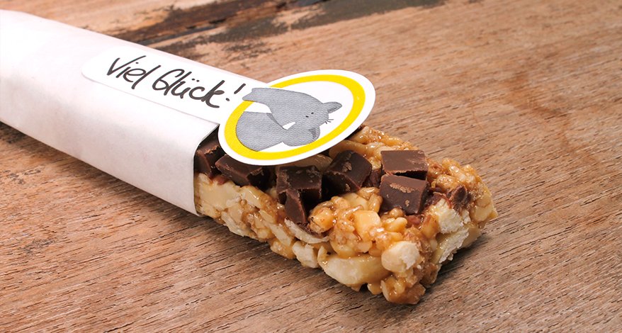 Ein Müsliriegel ist halb in Butterbrot-Papier eingepackt. Auf dem Butterbrot-Papier klebt ein Aufkleber der Marke Avery Zweckform Living für Kinder mit einem Elefanten-Motiv. Auf dem Aufkleber steht "Viel Glück".
