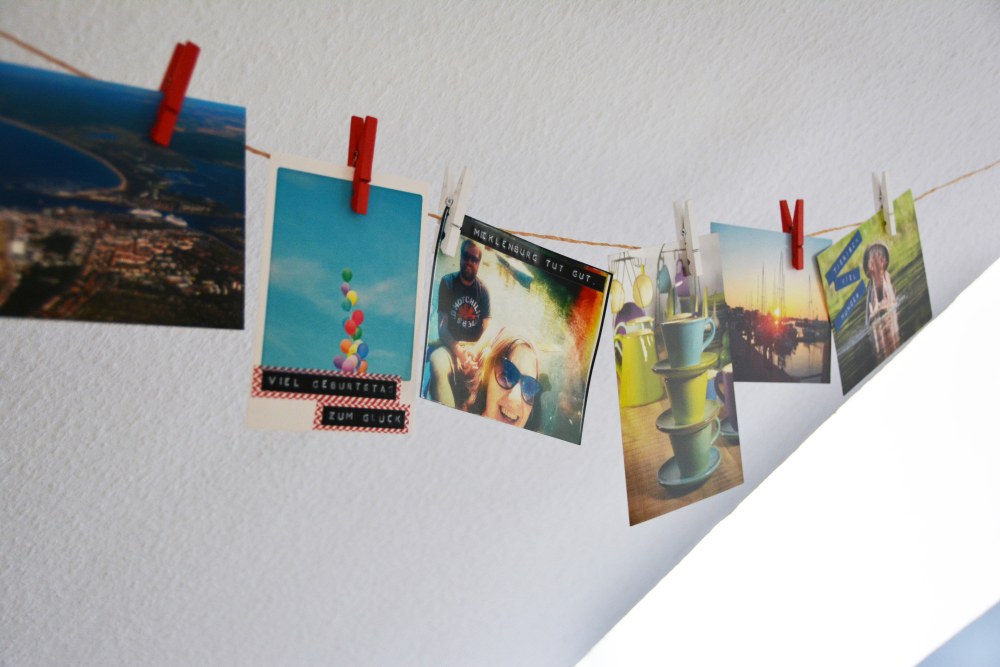 An einer weißen Wand läuft eine dunkle Kordel entlang, an die mit kleinen Klammern verschiedene Fotos gehängt wurden. Diese wurden mit Prägebändern von Dymo beschriftet.