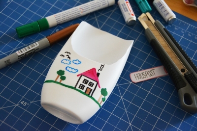 Auf einer Schneidematte liegt eine aufgeschnittene weiße Shampooflasche, auf die mit Permanentmarkern ein Haus mit Himmel und Wiese gemalt wurde. Daneben leigen bunte Permantenmarker und ein Cuttermesser.
