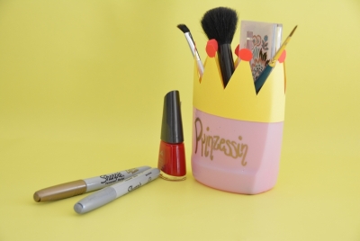 Aus einer leeren Shampooflasche wurde eine Aufbewahrung für Schminkutensilien gebastelt. Auf der rosa Flasche, die als Krone gestaltet ist, steht in gold "Prinzessin". Daneben liegen 2 Sharpie Stifte.