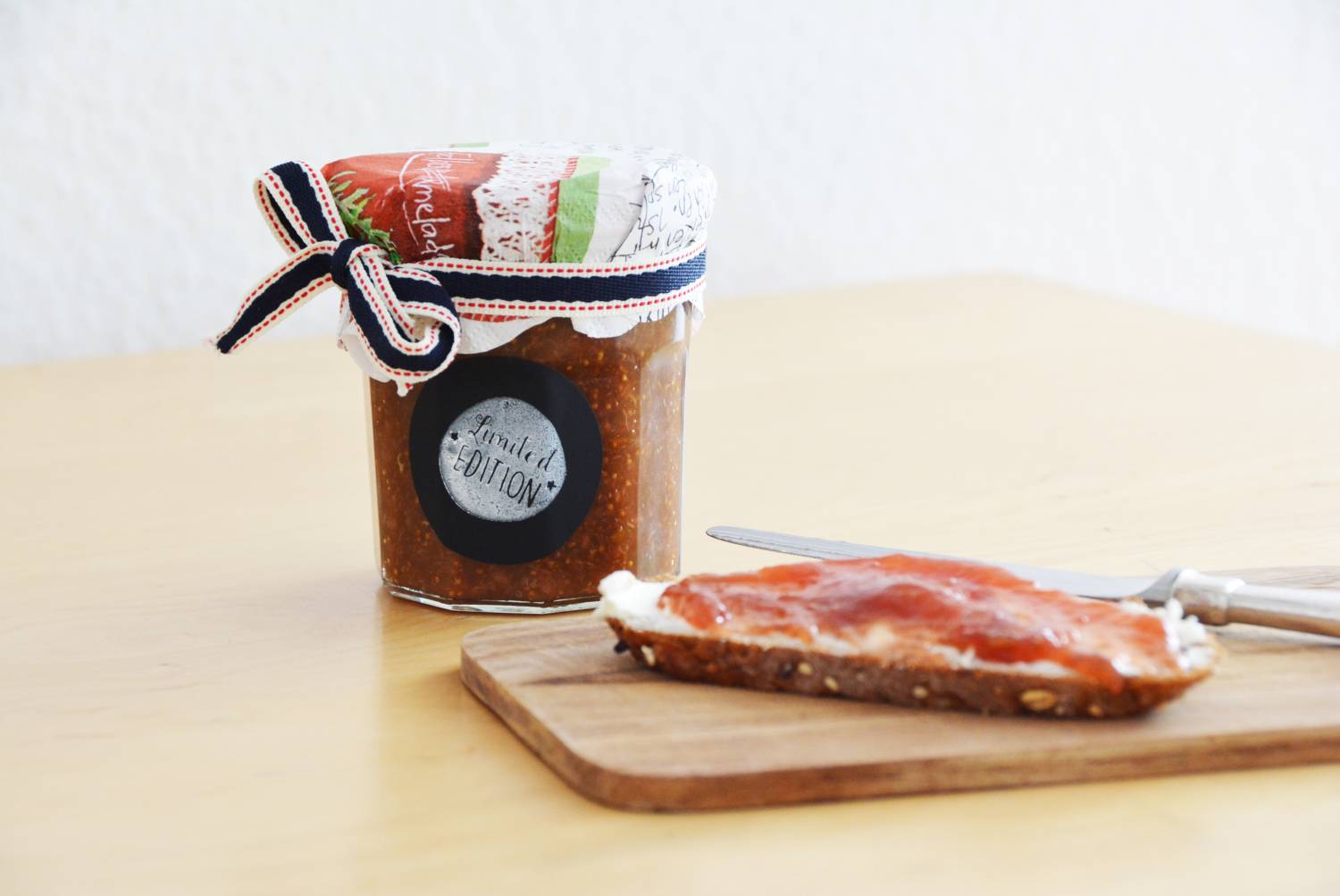 Auf einem Tisch liegt ein Frühstücksbrettchen, auf dem ein Marmeladenbrot liegt. Hinter dem Brettchen steht ein Glas mit Marmelade.