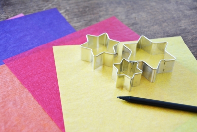 Auf einem Tisch liegen blaues, pinkes, oranges und gelbes Transparentpapier sowie 3 Stern Ausstecher in unterschiedlichen Größen und ein Bleistift.