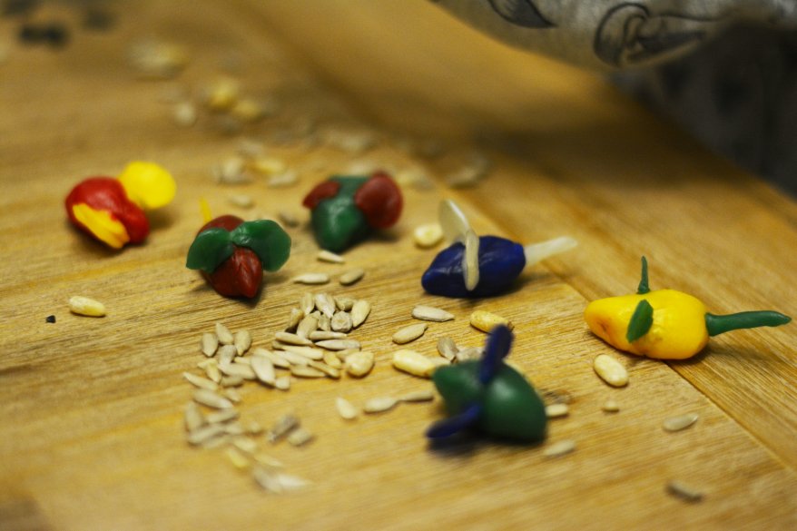 Auf einem Holztisch liegen, aus Knetbienenwachs geformte, bunte Mäuse. Dazwischen liegen Körner, die als Futter für die Mäuse dienen soll.