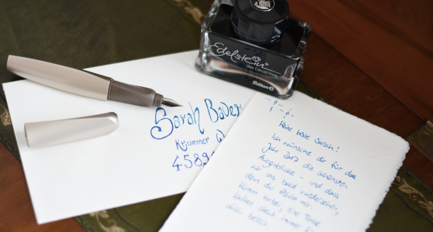 Auf einem Tisch liegt ein handgeschriebener Brief inklusive beschriftetem Briefumschlag. Auf dem Briefumschlag liegt ein Pelikan Twist Füller und ein Fass mit blauer Edelstein-Tinte der Marke Pelikan steht daneben.