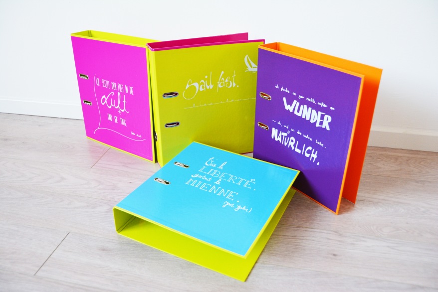 Auf dem Boden stehen mehrere bunte Ordner der Marke Pagna, welche mit Kreidemarker beschriftet und belettert sind.