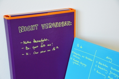 Auf einem lila Ordner ist mit Kreidemarker geschrieben: "Nicht vergessen: Mathe Hausaufgaben, Do.:Sport fällt aus, Mi.: Chor startet um 16 Uhr". Auf einer hellblauen Mappe sind 2 To-Do-Listen aufgeschrieben.
