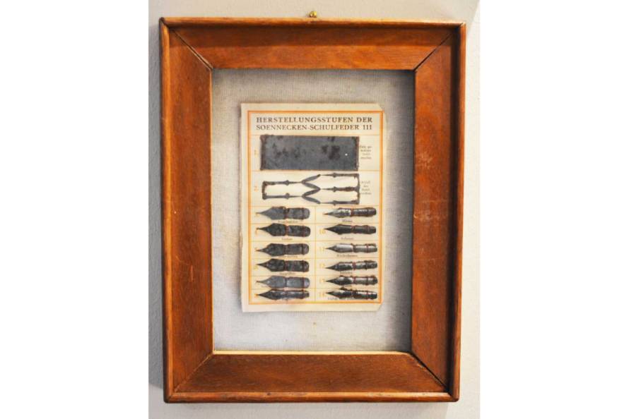 In einem braunen Holzbilderrahmen hängt ein Bild mit den Herstellungsstufen einer Soennecken Schulfeder.