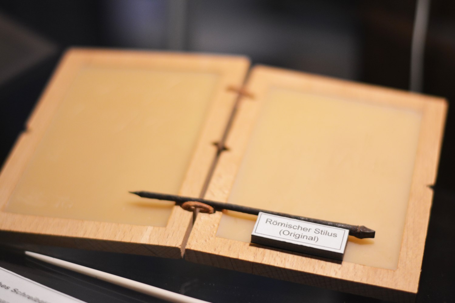 Auf einer Holztafel liegt ein römischer Stilus. Darunter sieht man das Hinweisschild mit der Erklärung "Römischer Stilus (Original)".