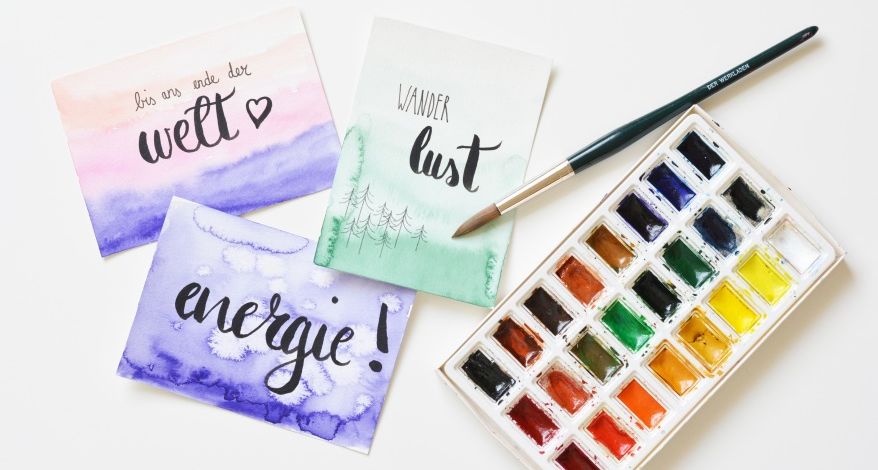 Flatlay mit geletterten Karten, auf denen "energie", "Wanderlust" und "bis ans Ende der Welt" steht. Daneben liegen ein Aquarell Farbkasten und ein Pinsel.