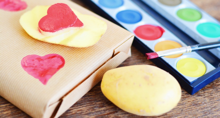 Eine Kartoffel mit einem roten Herz bemalt und auf ein eingepacktes Geschernk gedrückt. Daneben ein offener Wassermalkasten.