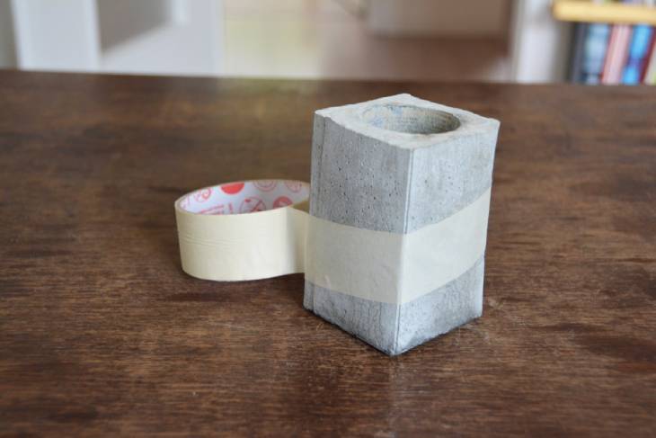 Auf einem dunbklen Holztisch steht der fertig ausgehärtete Betonbecher, welcher für eine weitere Verzierung mit Klebeband abgeklebt ist.