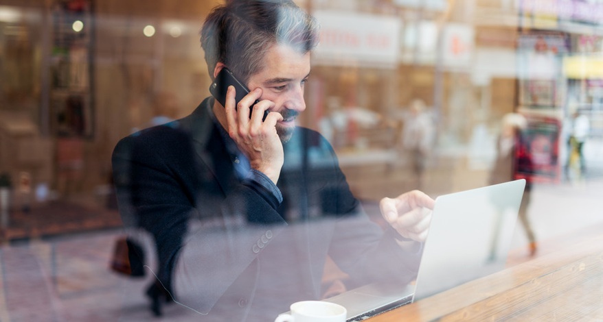 Ein Mann in Anzug sitzt in einem Café vor dem Fenster. Er telefoniert mit seinem Smartphone und hate seinen Laptop vor sich aufgeklappt.