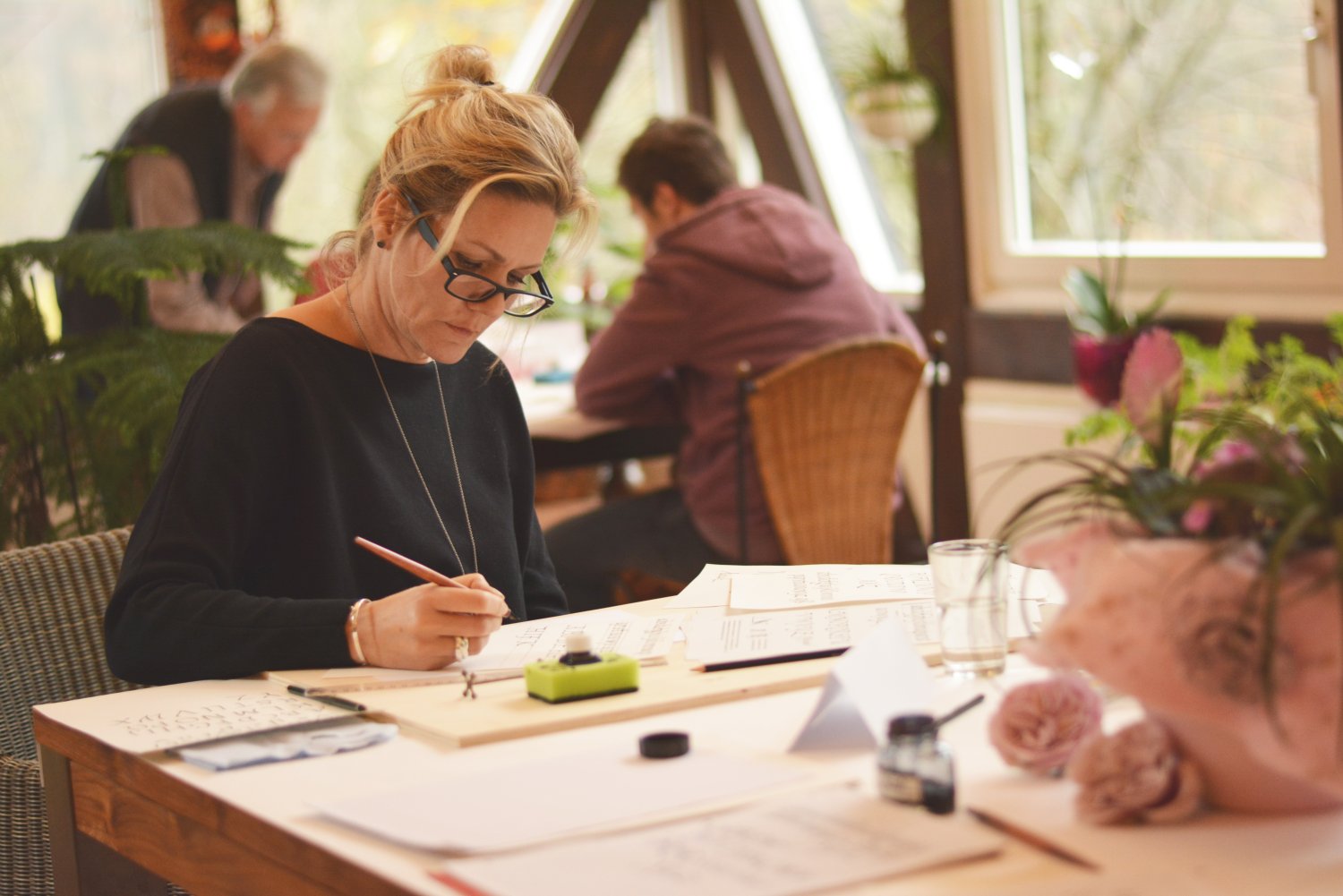 Eine Frau sitzt an einem Tisch. Vor ihr liegen viele Blätter mit Kalligrafie. Sie hält eine Schreibfeder in der Han, vor ihr steht ein Tintenfass.