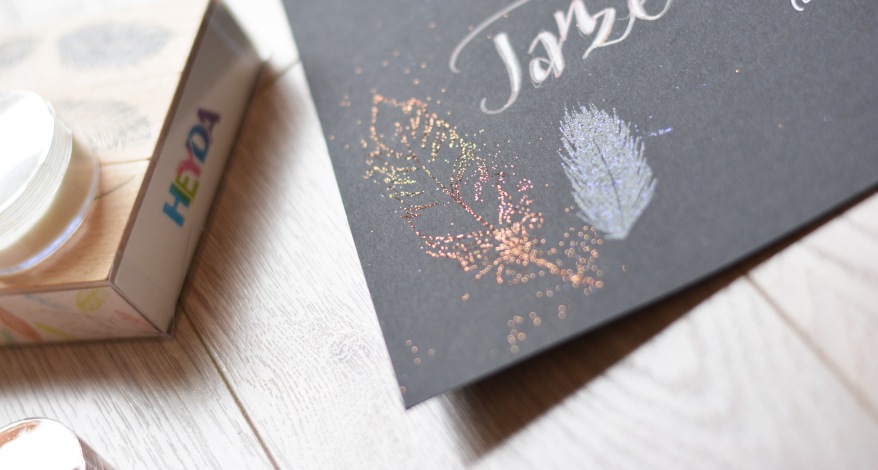 Schwarzes Papier mit schimmernden Federstempeln und Kalligrafie „Tanze…“, daneben ein Stempel von Heyda.