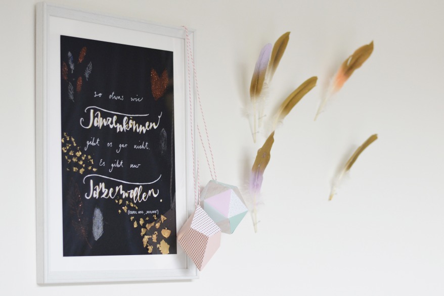 Gerahmtes Zitat mit Golddeko und Federn an der Wand, daneben Dekoobjekte.