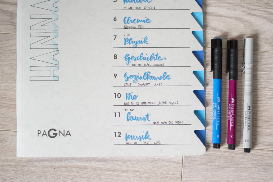 Eine Ordnungsmappe von pagna wurde mit Handlettering gestaltet. Daneben liegen drei Fineliner und Handletteringstifte von Faber-Castell.