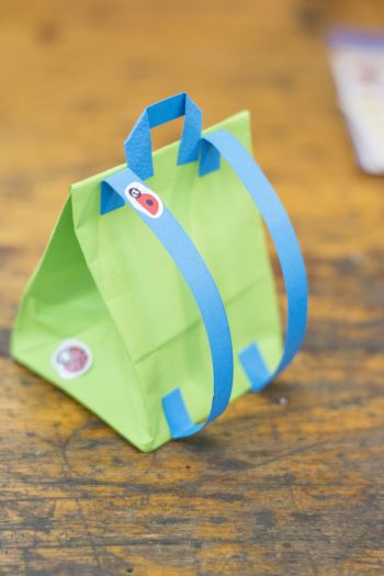 Eine Geschenktüte aus grünem Papier in Form eines Schulranzens steht auf einem alten Holzboden.. Die Schulteriemen und der Tragegriff sind aus blauen Papier gefertigt. Die Tüte wurde mit Marienkäfer-Stickern beklebt.