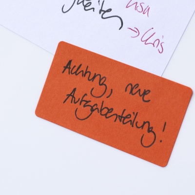 Nahaufnahme eines orangen Etiketts mit der Aufschrift "Achtung, neue Aufgabenteilung!"