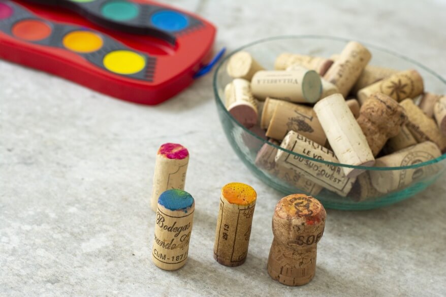 Weinkorken mit bunten Farben neben einer Schale weiterer Korken und einem Farbkasten.