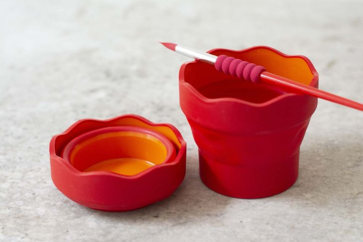 Zwei rote Wasserbecher von Faber-Castell mit einem roten Pinsel auf grauem Untergrund.
