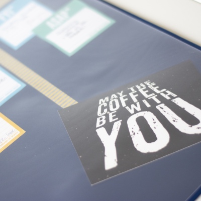 In einer Schreibtischunterlage der Marke Durable ist eine Postkarte befestigt. Diese hat den Text "May the Coffee be with you"