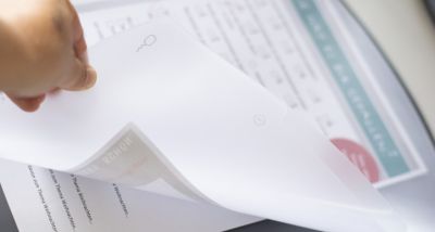 Eine Hand hält die Abdeckung einer Schreibtischunterlage hoch. Darunter befinden sich diverse Zettel. 