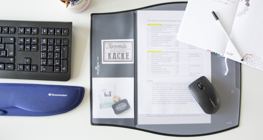 Auf einem weißen Schreibtisch sind eine schwarze Tastatur, eine Handauflage der Marke Soennecken sowie eine Schreibtischunterlage von Durable zu sehen. Auf der Unterlage liegt eine Maus und ein aufgeschlagenes Notizbuch, in dem ein Fineliner liegt. In der Schreibtischunterlage sind verschiedene Zettel und Aufkleber aufbewahrt.