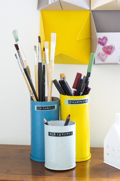 Drei DIY-Stiftehalter aus bunt lackierten Dosen gefüllt mit Pinseln und Stiften.