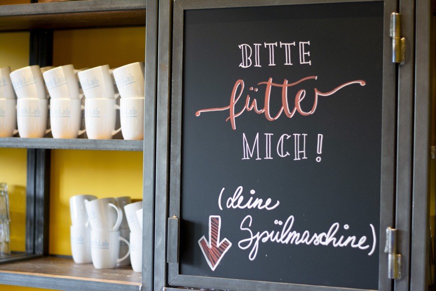 Tafel mit Aufschrift „Bitte füttere mich“ über Geschirrspüler.