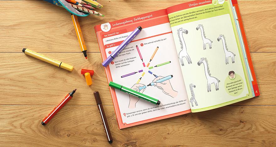 Offenes Übungsbuch mit bunten Stiften auf einem Holztisch.