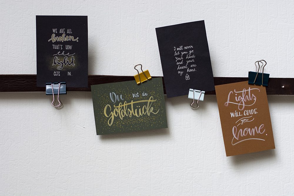 Vier Lettering-Karten mit Sprüchen in Gold und Weiß, aufgehängt mit Büroklammern.