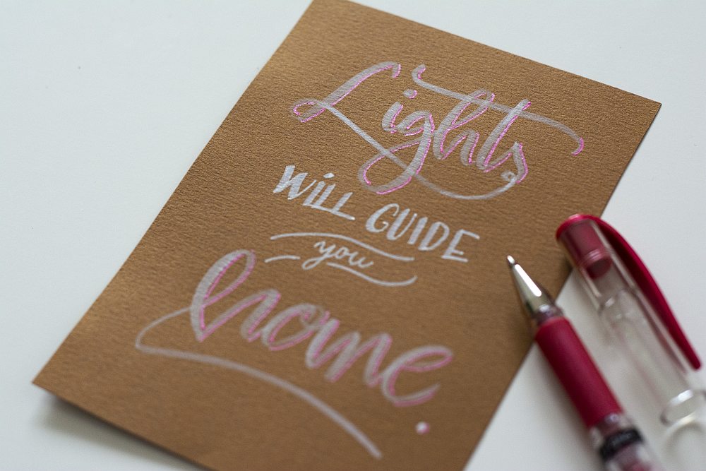 Handlettering „Lights will guide you home“ in silber und rosa glänzend auf brauner Karte.